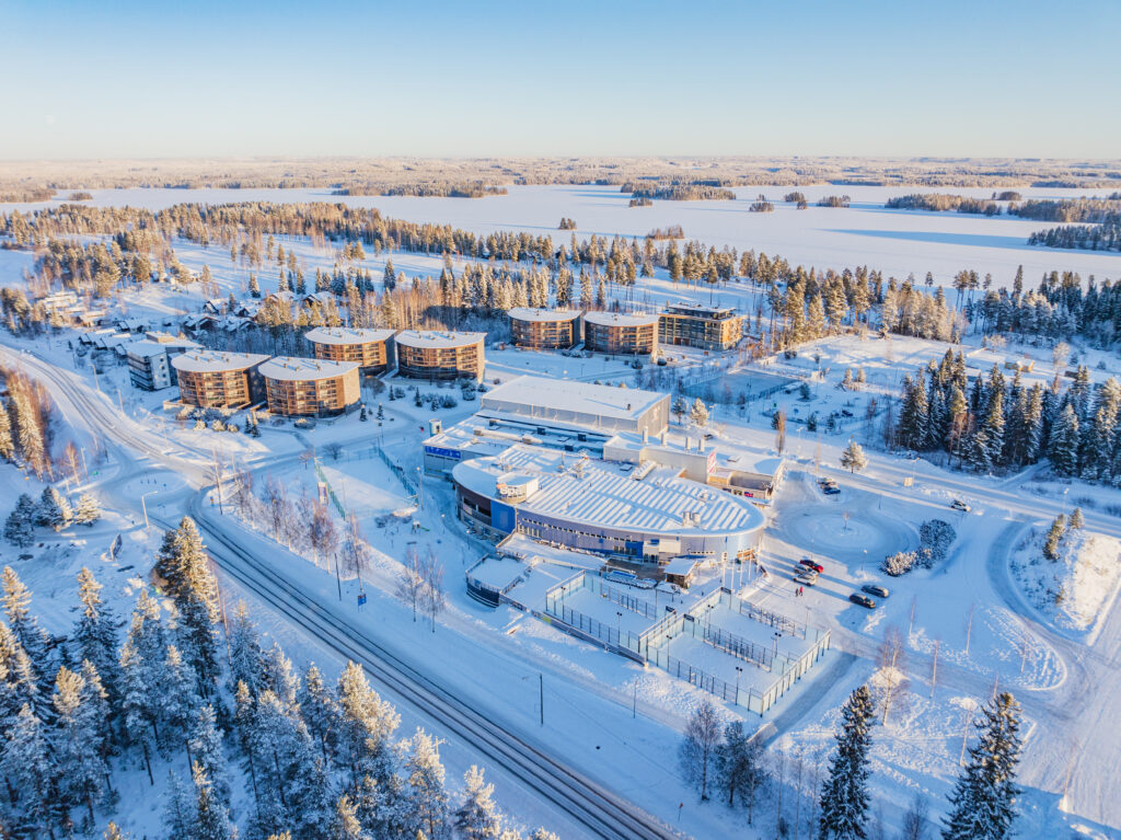 Ilmakuva talvisesta Tahkon keskustasta. Matalalta paistava aurinko valaisee lumiset tiet, metsän ja hotellirakennuksen. Horisontti näkyy kaukana taustalla.